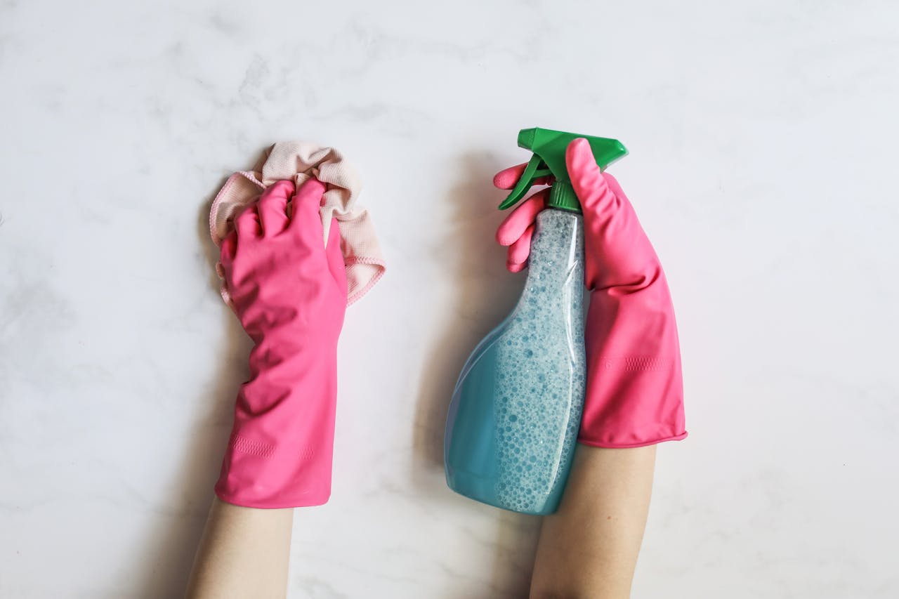 about-01 A pair of hands in pink gloves are cleaning with a spray bottle on a white background.